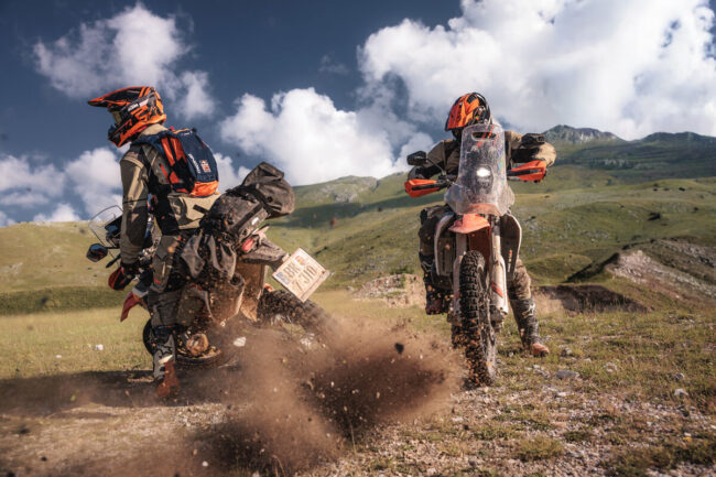 Ujęcie akcji, w którym KTM 690 RALLY 2027 pokonuje ciasny, techniczny zakręt na podłożu mieszanym (piach i luźne kamienie). Motocykl jest mocno pochylony, a kierowca demonstruje lekkość i zwinność konstrukcji LC4, dynamicznie wyprowadzając maszynę z łuku. Widać pracujące zawieszenie WP XPLOR oraz jak opony Continental TKC 80 wgryzają się w nawierzchnię, zapewniając trakcję. Tło jest lekko rozmyte, co dodatkowo podkreśla dynamikę ruchu i precyzję prowadzenia, udowadniając, że duży zasięg nie oznacza kompromisów w trudnym terenie.