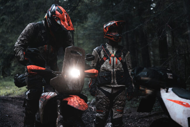 KTM 690 RALLY 2027 zaparkowany na szczycie wzgórza, z którego roztacza się panoramiczny widok na rozległą dolinę i krętą, szutrową drogę prowadzącą w nieznane. Motocykl, zwrócony przodem do zapierającego dech w piersiach krajobrazu, jest oświetlony ciepłymi promieniami słońca. Ten kadr symbolizuje obietnicę większego zasięgu (dzięki 20 litrom paliwa) i gotowość do długodystansowego adventure'owania. Wyświetlacz TFT jest czytelny, a rajdowa sylwetka motocykla wyraźnie odcina się od surowego, górskiego tła, zachęcając do eksploracji.