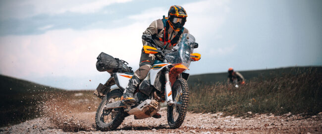KTM 690 RALLY 2027 ujęty w dynamicznym kadrze z profilu, podczas jazdy w zróżnicowanym, szutrowo-kamienistym terenie. Motocykl, w charakterystycznym, rajdowym malowaniu KTM, znajduje się w lekkim przechyle, a przednie koło precyzyjnie omija przeszkodę. Kierowca w pełnym rynsztunku, stojąc na szerokich podnóżkach, idealnie balansuje ciałem. Doskonale widoczna jest rajdowa wieża ze zintegrowaną szybą oraz oba przednie zbiorniki paliwa, które nadają maszynie potężny, a jednocześnie zwarty wygląd, sugerując jej pełną gotowość do długich etapów.